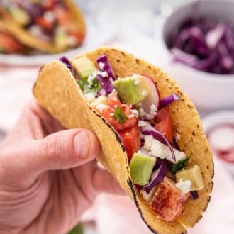 Pinterest graphic of a hand holding up a lobster taco with tomatoes, avocado, radishes, and cabbage as a topping.
