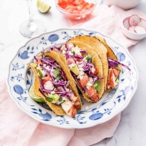 A plate of three lobster tacos on corn tortillas on top of a pink linen napkin with a lime wedge, bowl of sliced radishes, and bowl of diced tomatoes in the background.