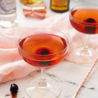 Pinterest graphic of two glasses of Manhattans, each with a maraschino cherry in the middle of each and one on the counter top beside a pink linen napkin.