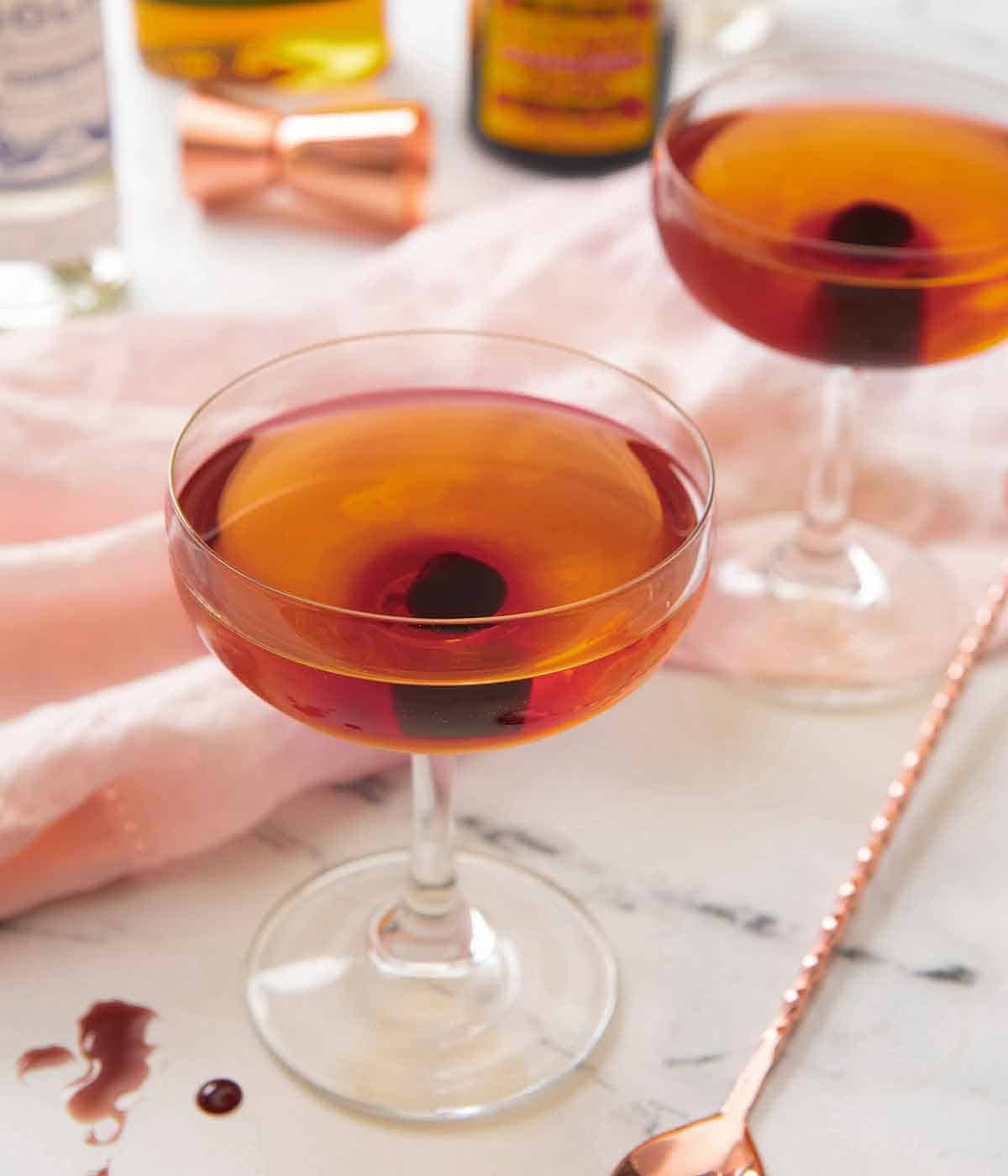 Two glasses of Manhattan cocktails with a maraschino cherry in each. Both drinks are in front of a pink linen napkin with a cocktail stirrer.