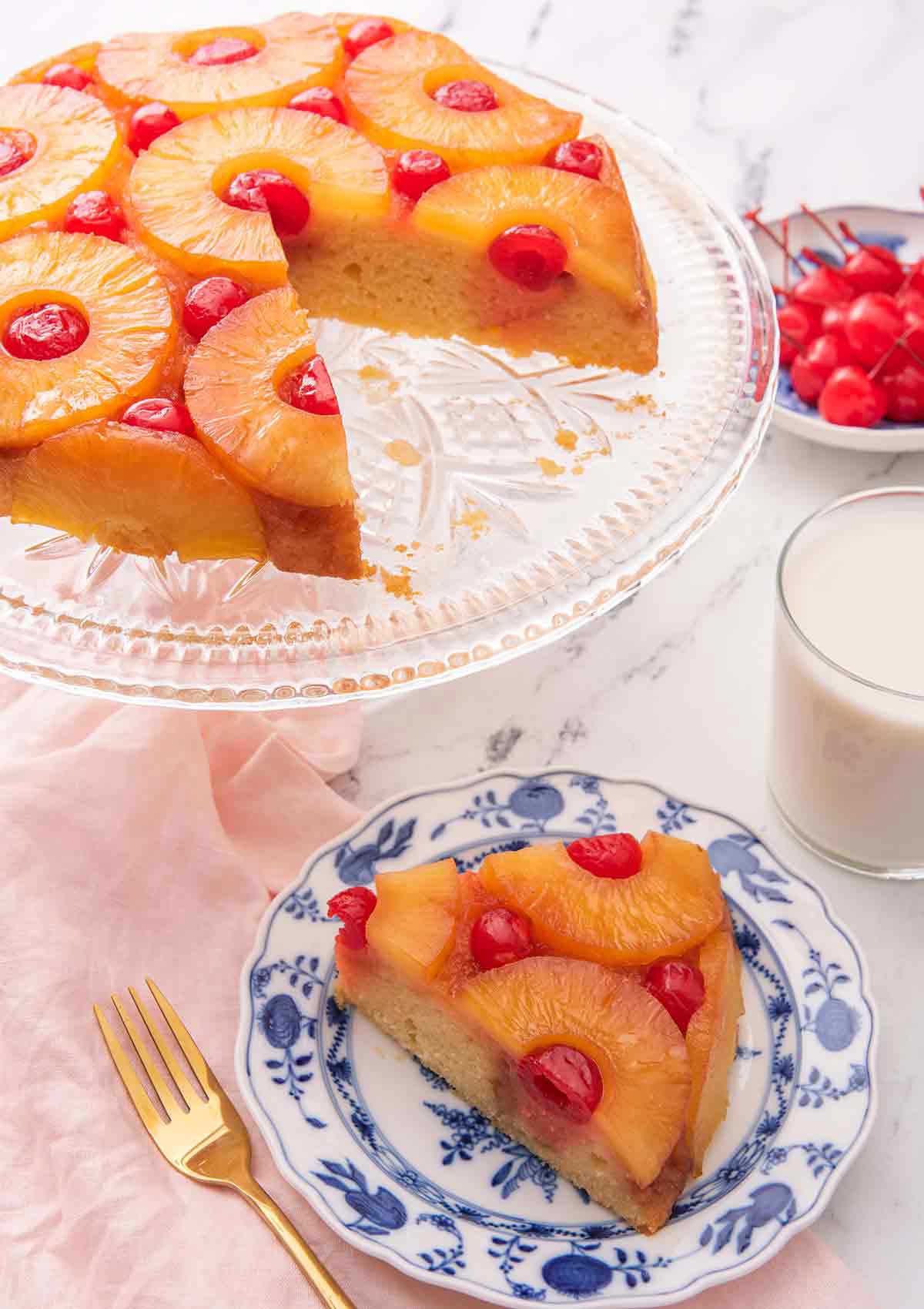 A pineapple upside down cake on a cake stand with a slice cut out and placed on a plate in front of it.