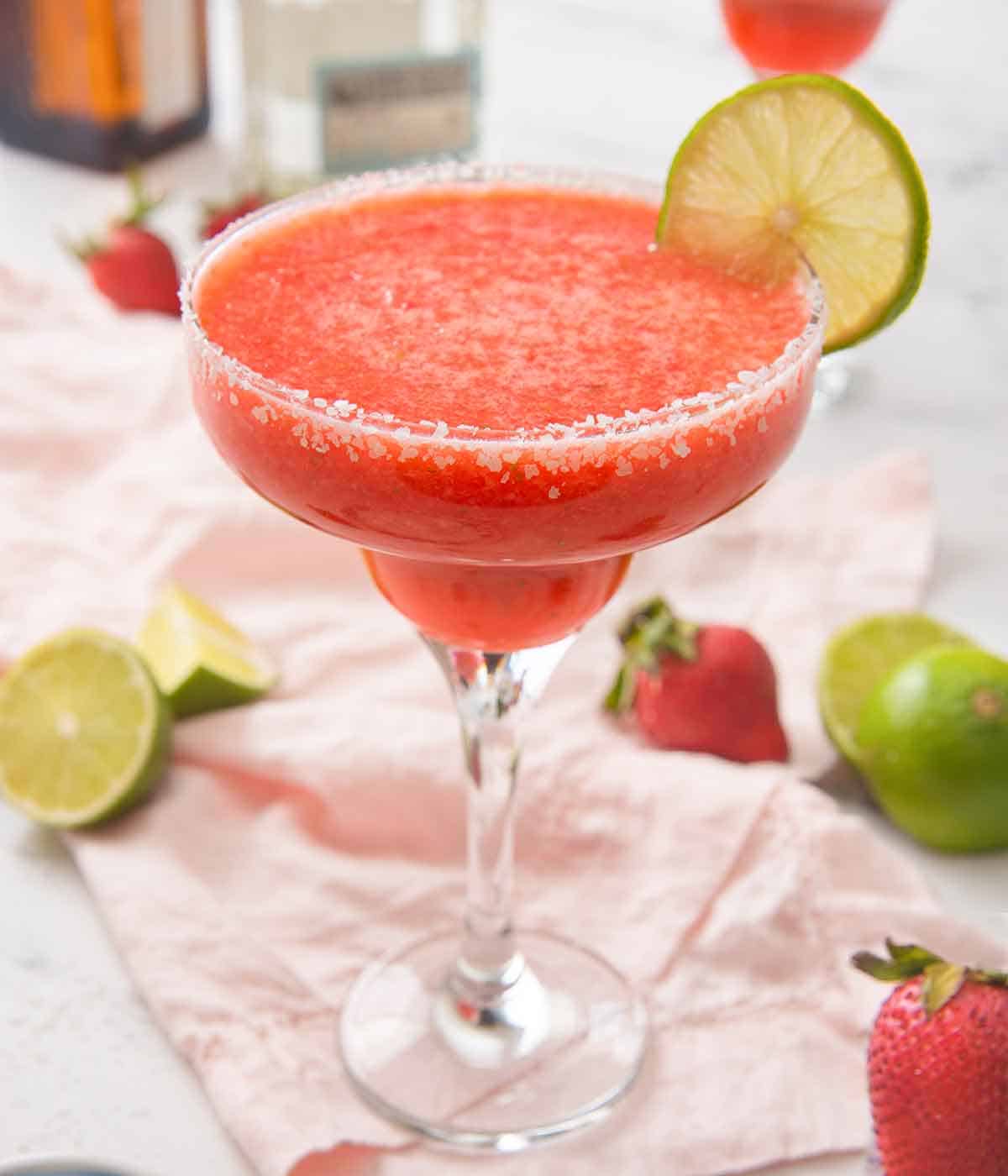 A cocktail glass filled with a pink strawberry margarita with a lime slice on the rim on top of a pink linen napkin with cut limes and strawberries scattered around.