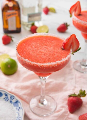 A strawberry margarita in a cocktail glass with a halved strawberry on the rim with the ingredients out of focus in the background.