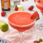 Close up of a glass of strawberry margarita with a strawberry on the rim. Ingredients scattered in the background out of focus behind the drink.