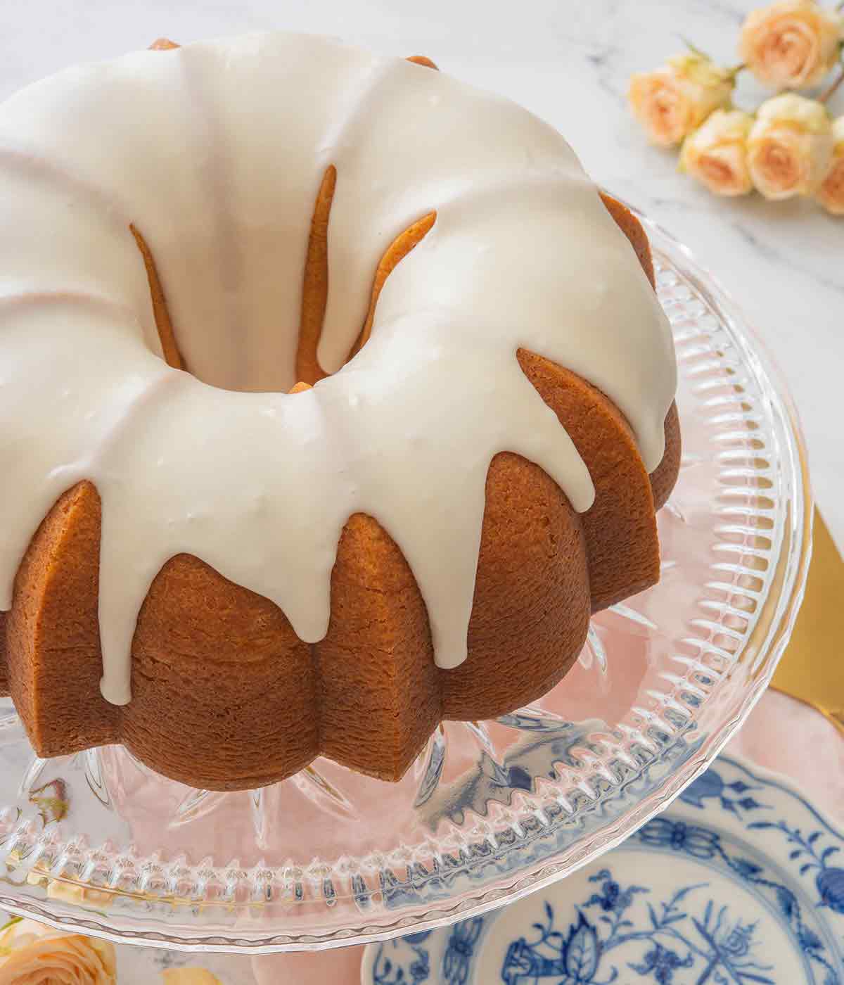 Close up of a glazed vanilla Bundt cake on a clear cake stand with roses in the background.