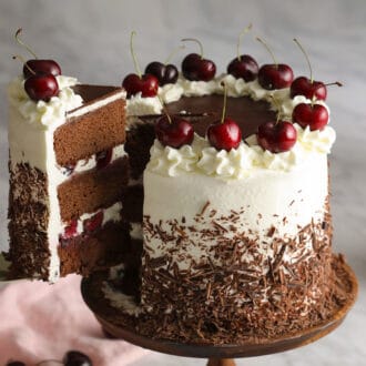 A black forest cake topped with cherries and covered in chocolate on a wooden cake stand.