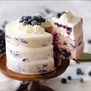 A blueberry lemon cake on a wooden cake stand with a piece getting removed