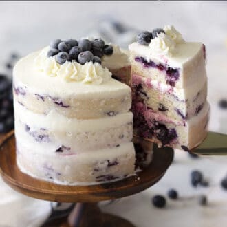 A blueberry lemon cake on a wooden cake stand with a piece getting removed