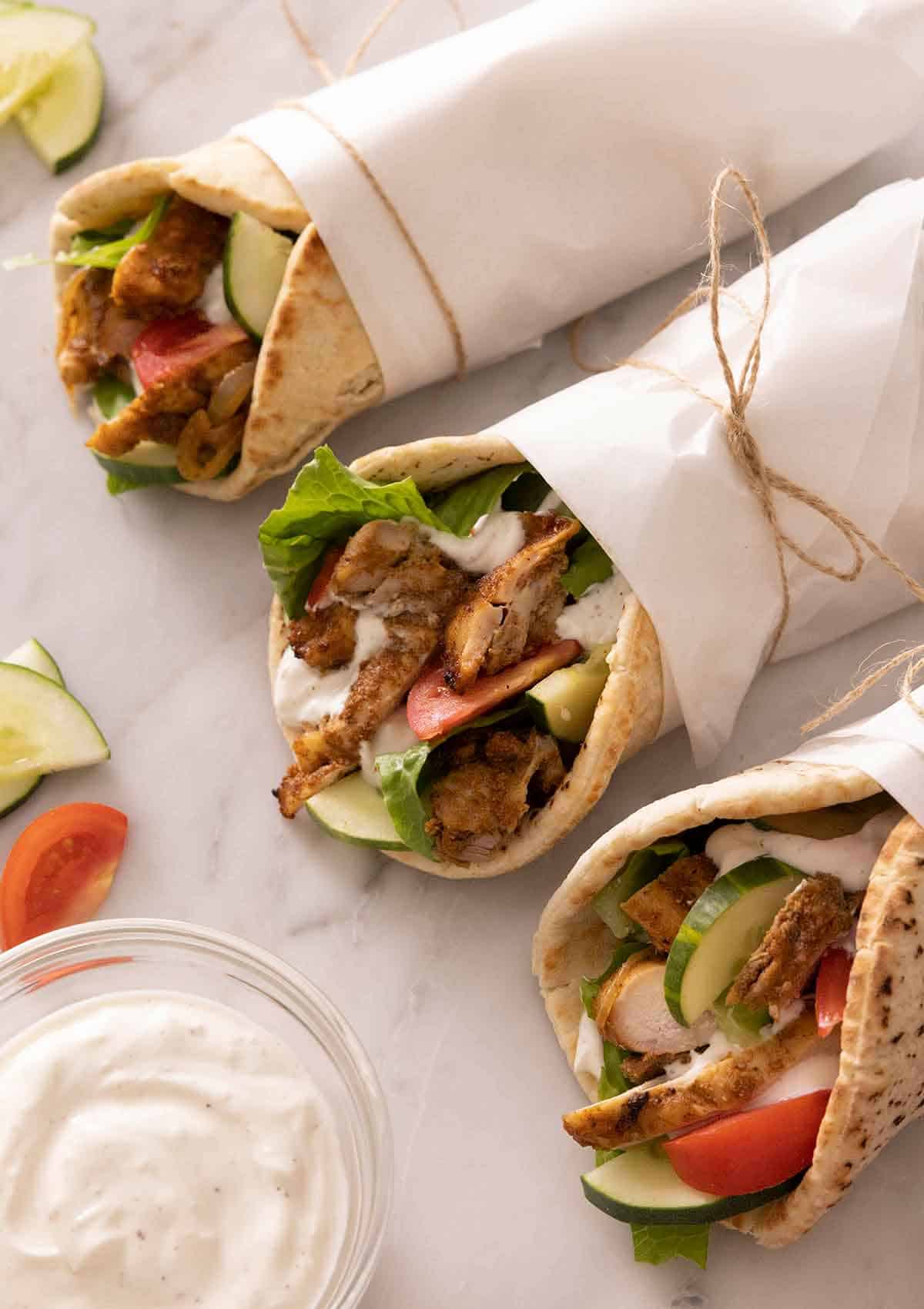 Three parchment wrapped chicken shawarma with a yogurt sauce on the side.