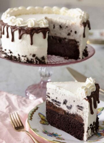 An ice cream cake on a cake stand with a slice cut out, on a plate in front.