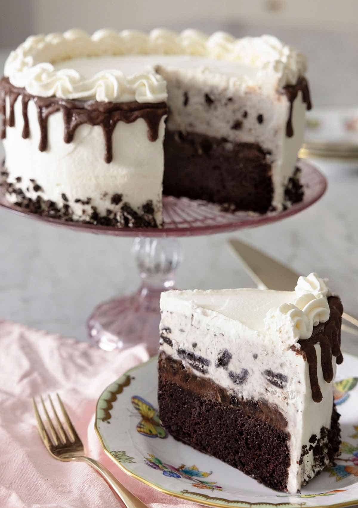 An ice cream cake on a cake stand with a slice cut out, on a plate in front.