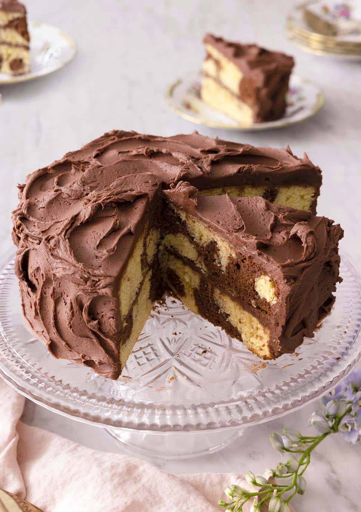 A cake stand with a chocolate frosted marble cake, with a quarter cut out. One cut slice is still on the stand and the other two are in the background on a plate.