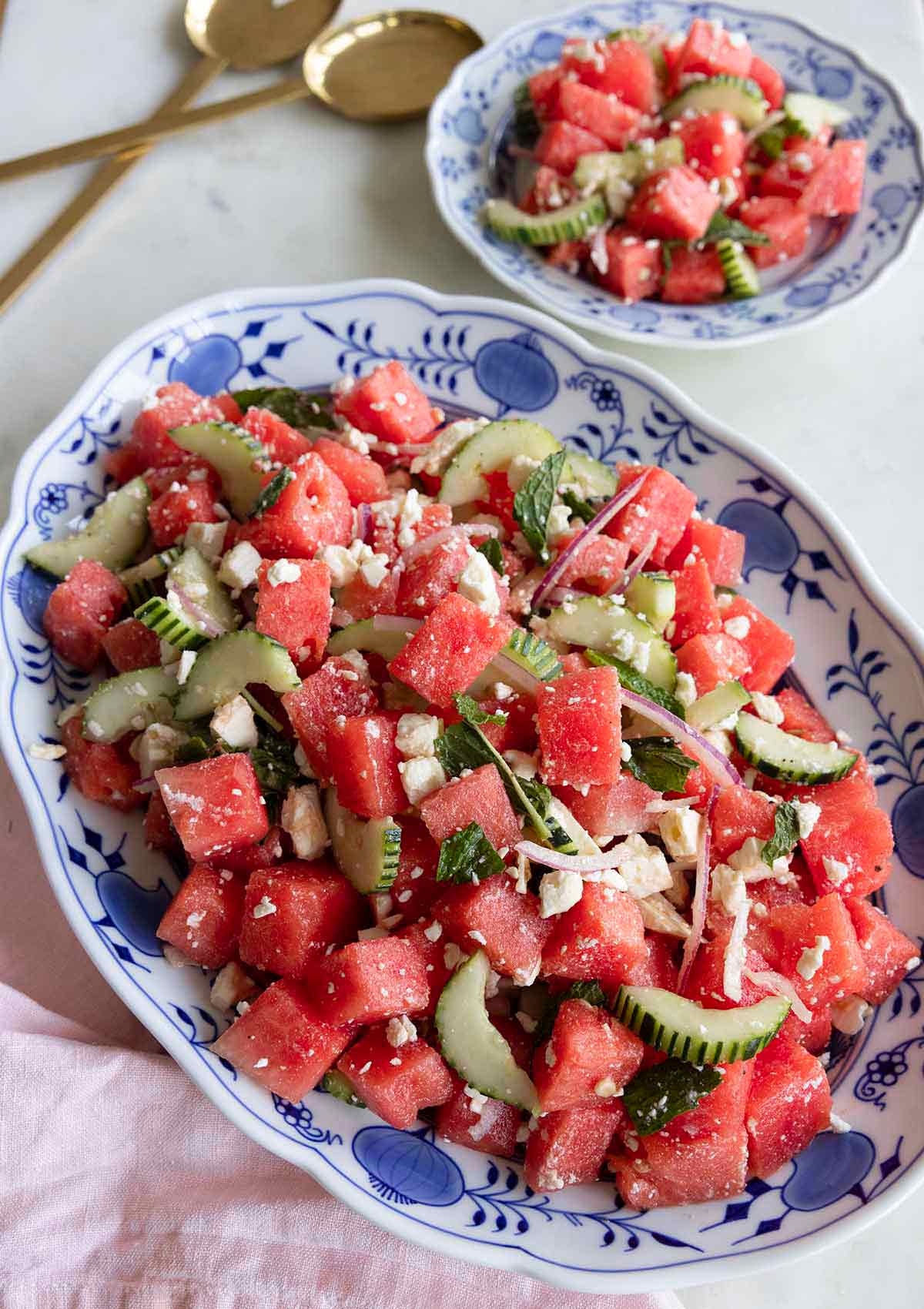 An oval platter of watermelon salad with a plate of it beside it along with serving spoons.