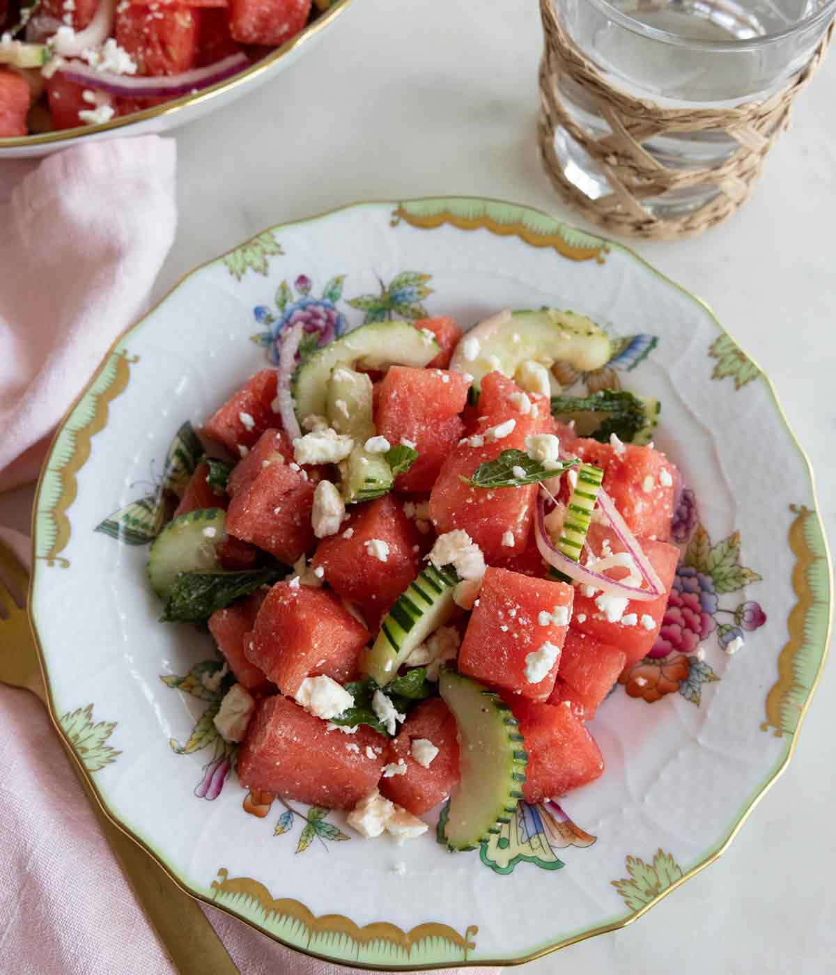 Overhead view of a plate of watermelon salad with a glass of water and a pink linen napkin beside it.