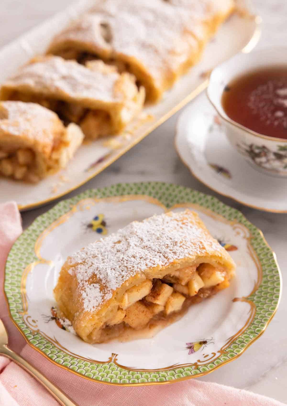 A serving of apple strudel on a plate in front of a cup of tea and more sliced apple strudel pieces.