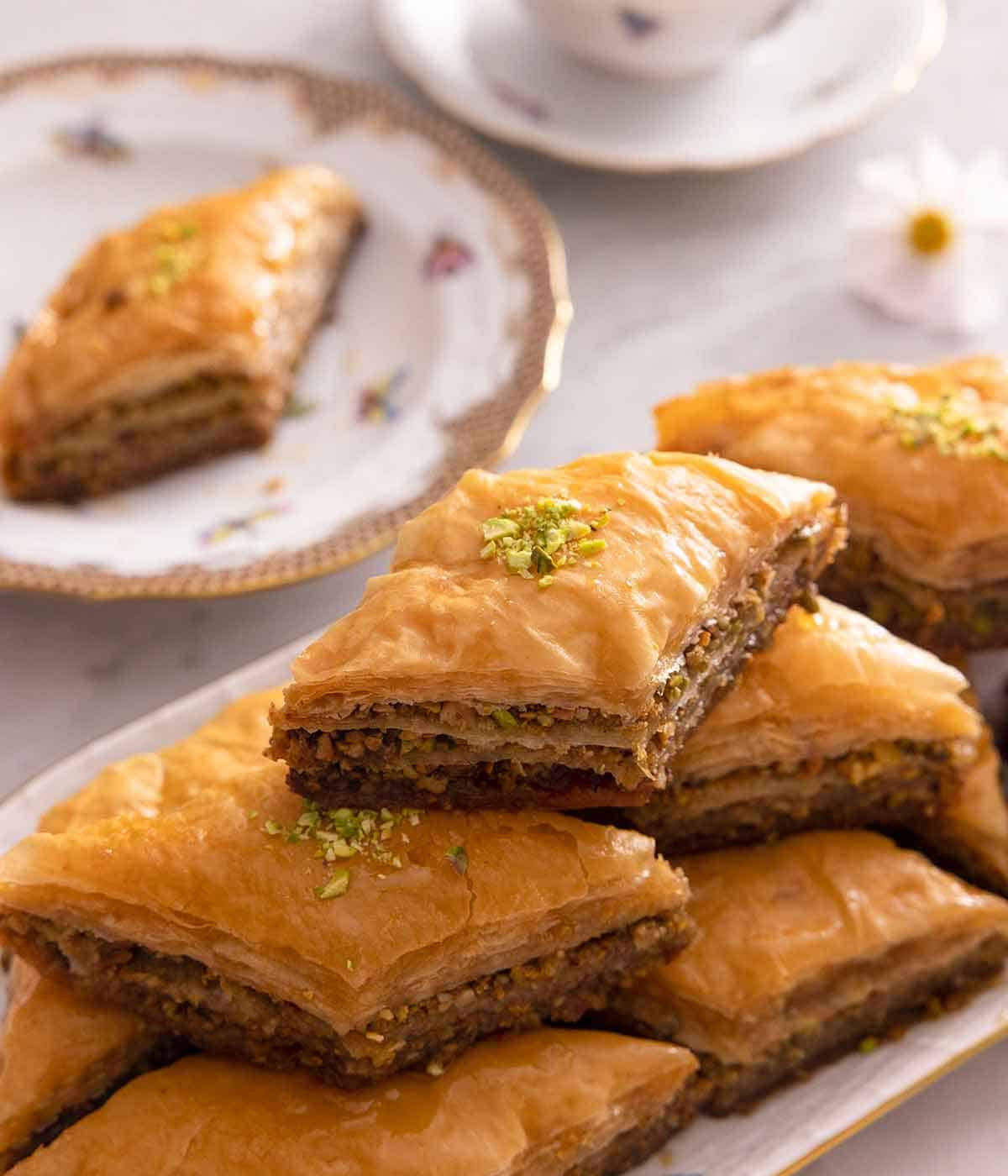 A platter of baklava stacked on top of each other with a plate with a single serving in the background.