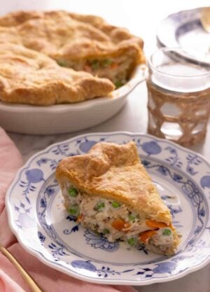 A plate of with a slice of chicken pot pie in front of a glass of water and the pie dish.