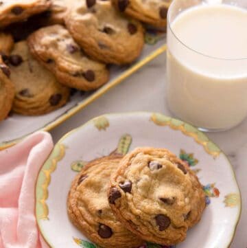 A plate with two chocolate chip cookies in front of a cup of milk and platter of cookies.