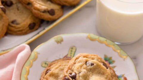 A plate with two chocolate chip cookies in front of a cup of milk and platter of cookies.