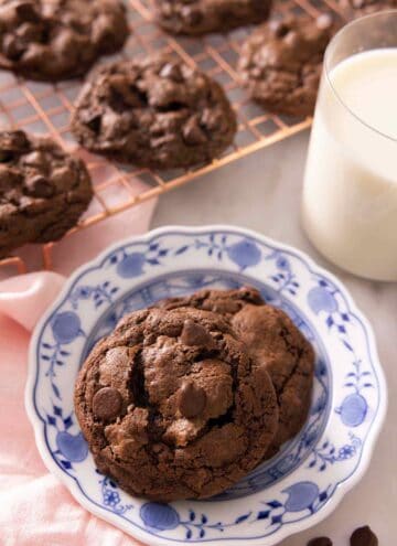 Two double chocolate chip cookies on a plate by a glass of milk with a cooling rack with more cookies in the background.