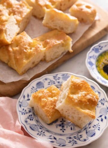 A plate with those pieces of focaccia in front of a cutting board with more pieces.