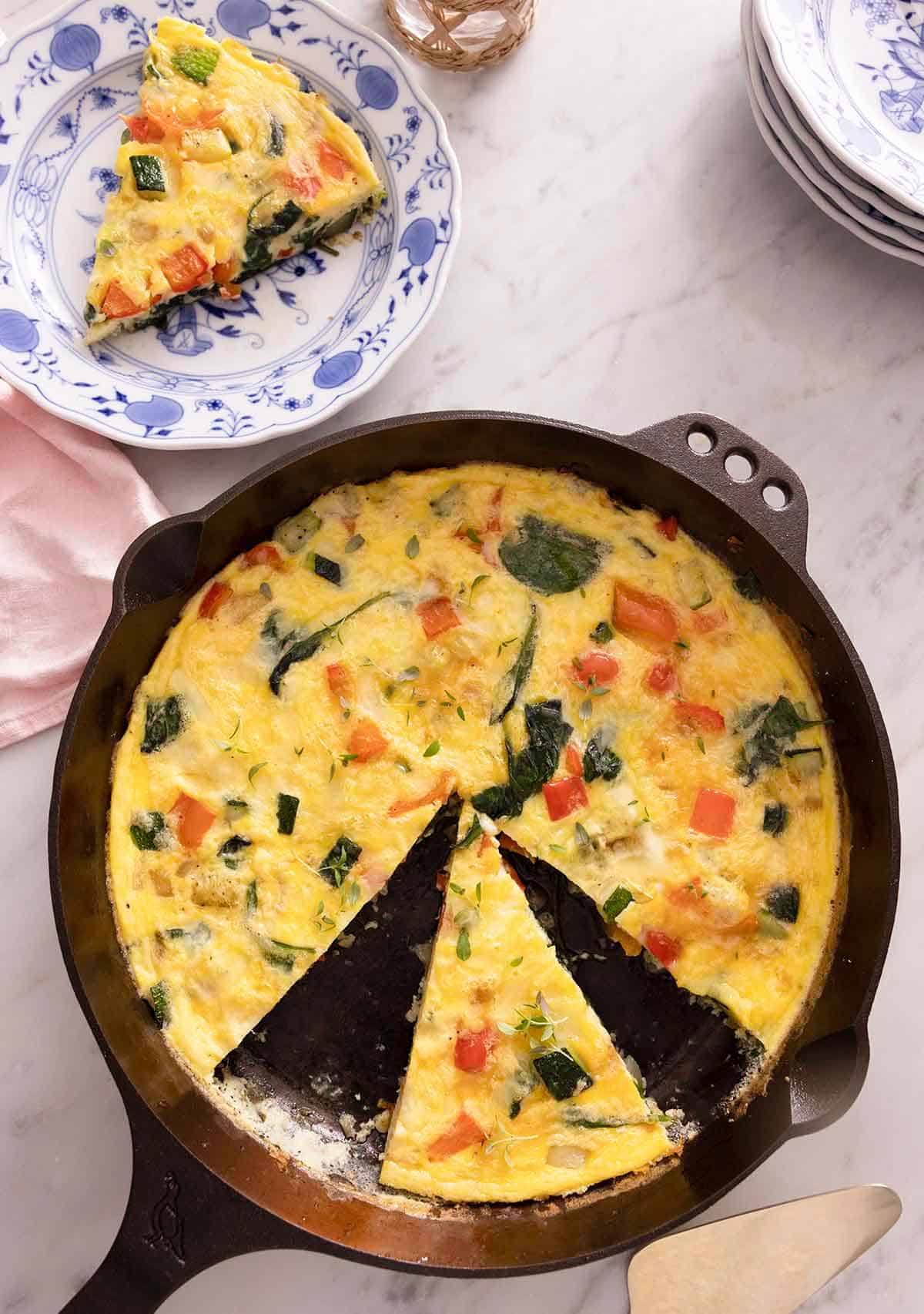 Overhead view of a pan with a frittata, cut, with a slice in a plate beside the pan.