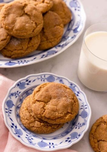 A plate with two pumpkin snickerdoodles with a larger plate containing more behind it with a glass of milk.