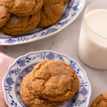 A plate with two pumpkin snickerdoodles with a larger plate containing more behind it with a glass of milk.