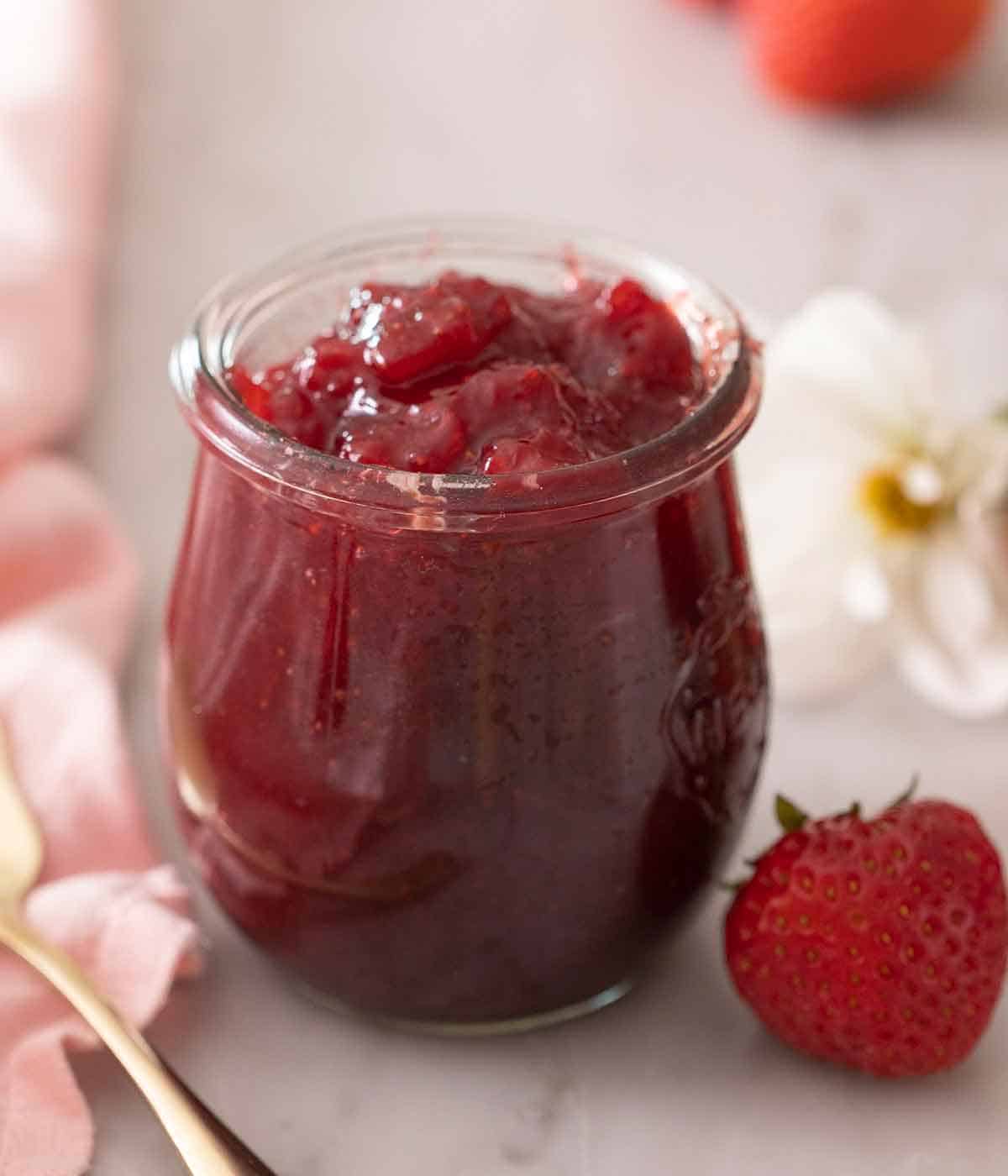 A tulip jar with strawberry jam with a strawberry in front.