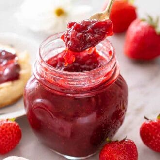 A mason jar with a spoon lifting the jam up in front of an english muffin on a plate with strawberries scattered around.