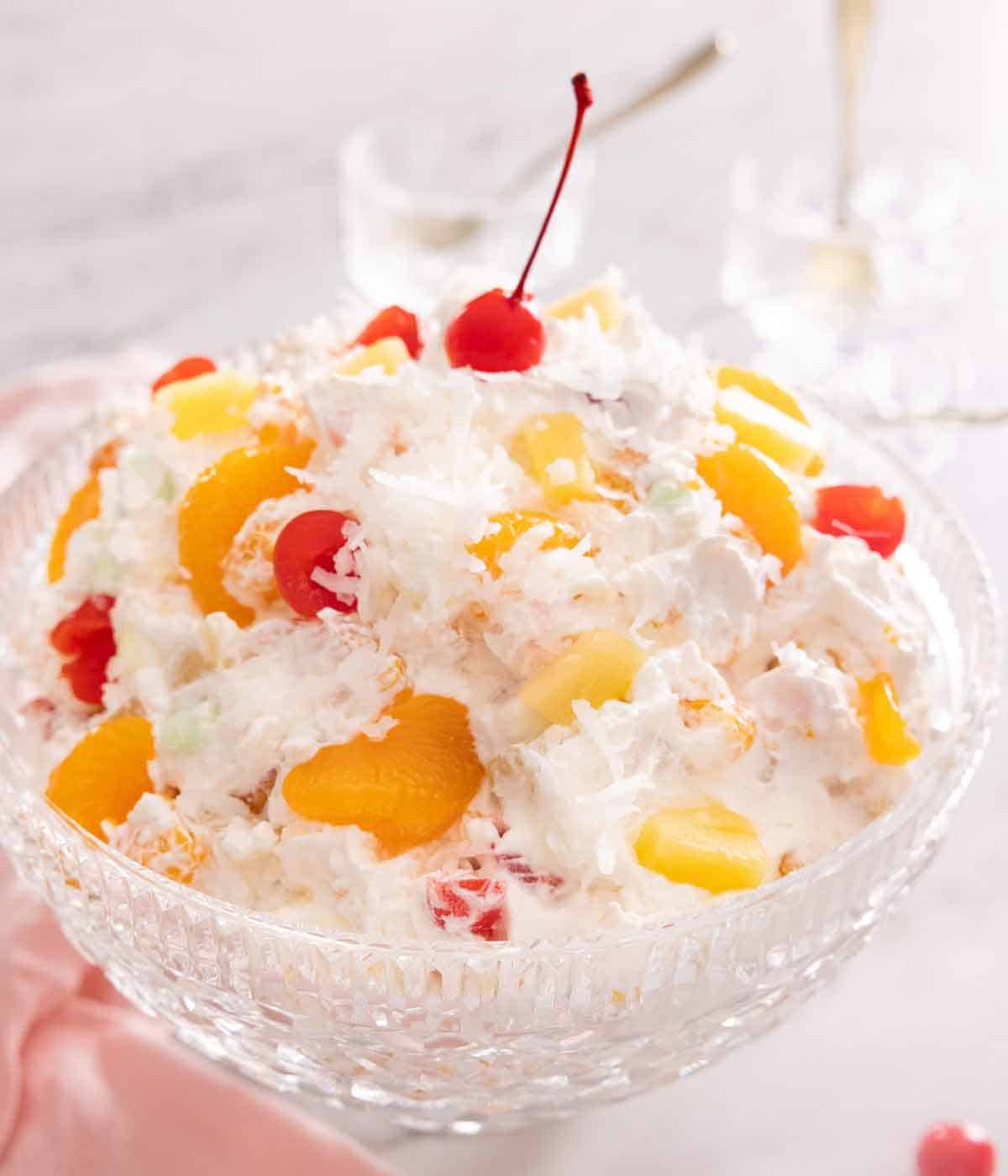 A serving bowl of ambrosia salad with fruit, coconut flakes, and whipped cream dressing.