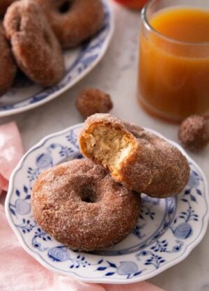 Two apple cider donuts, with one with a bite taken out of it, on a plate in front of a cup of apple cider.