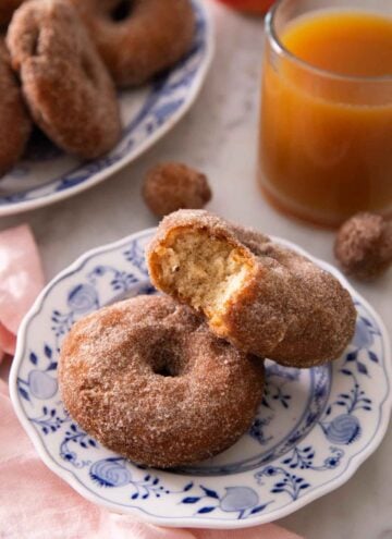 Two apple cider donuts, with one with a bite taken out of it, on a plate in front of a cup of apple cider.