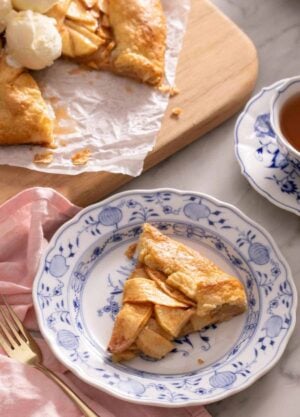 A plate with a slice of apple galette with a cutting board in the background with the rest of the galette along with a cup of tea.