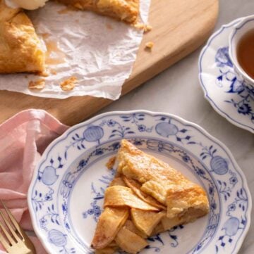 A plate with a slice of apple galette with a cutting board in the background with the rest of the galette along with a cup of tea.