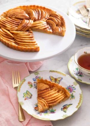A serving of apple tart in a plate in front of the entire tart that it was cut out of.