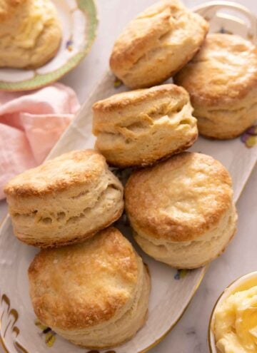 A platter with six biscuits with a plate off the corner with one biscuit and a bowl of butter in the other corner.