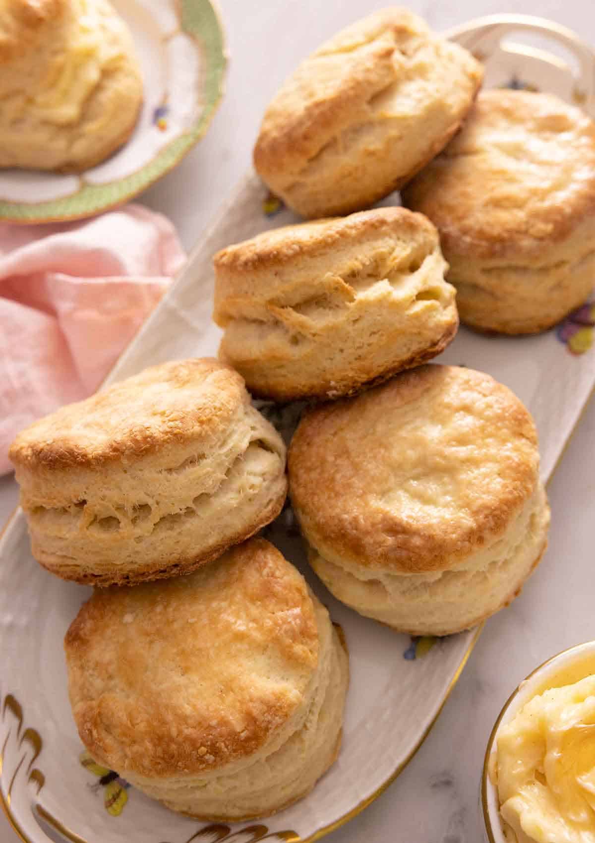 A platter with six biscuits with a plate off the corner with one biscuit and a bowl of butter in the other corner.