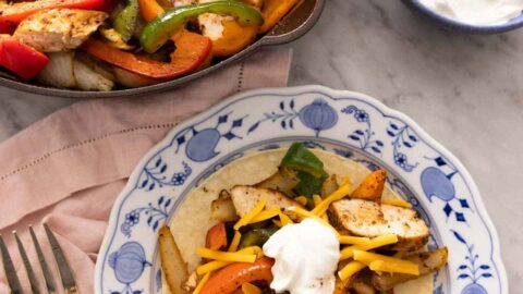 A plate with chicken fajitas on a tortilla in front of a pan of fajitas.