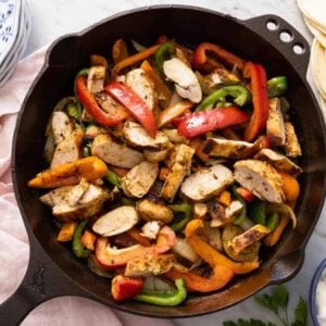 A cast iron skillet containing chicken fajitas beside a pink linen napkin, a bowl of sour cream, tortilla, and a stack of plates.