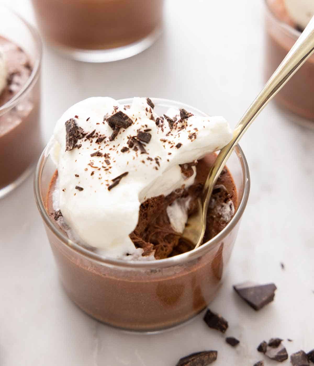 A spoon inside of a glass of chocolate mousse with whipped cream and chocolate. Small pieces of chocolate is scattered by the glass.