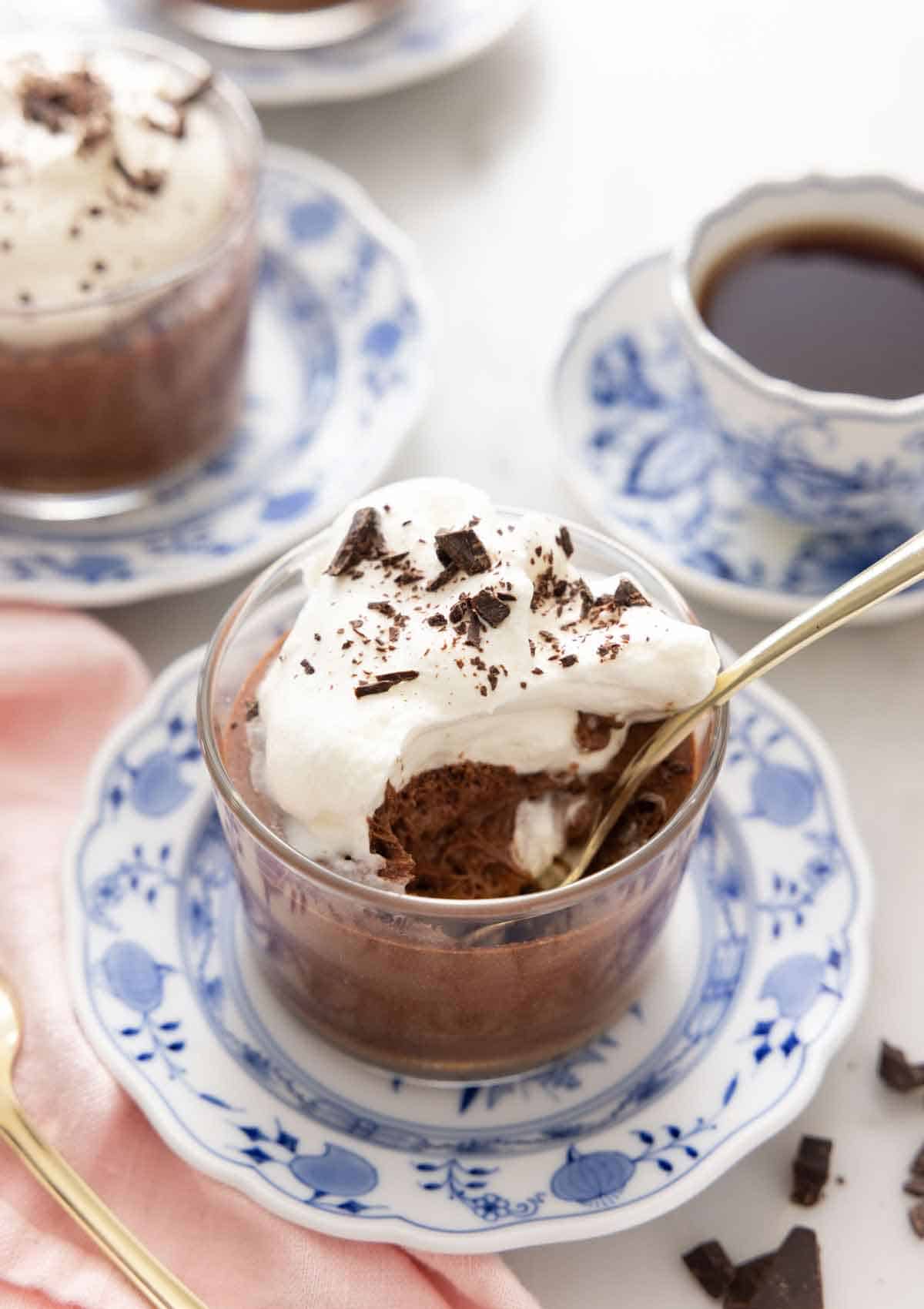 A glass of chocolate mousse with whipped cream and shaved chocolate with a spoonful eaten and a spoon inside.