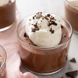 Four glasses of chocolate mousse with one in focus, in the middle, with whipped cream and shaved chocolate on top of all of them.