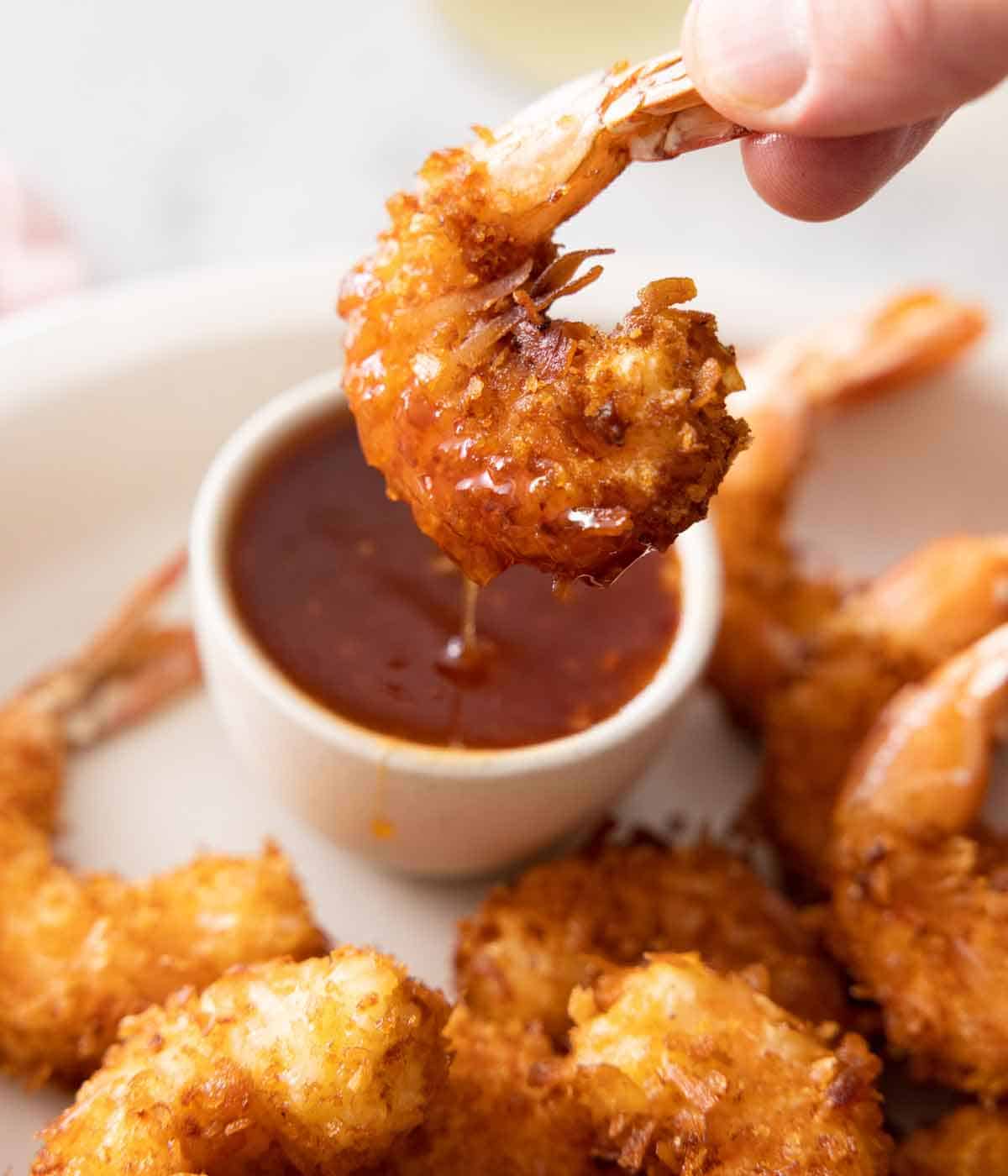 A coconut shrimp dipped into a bowl of sauce and lifted, with some sauce dipping off.