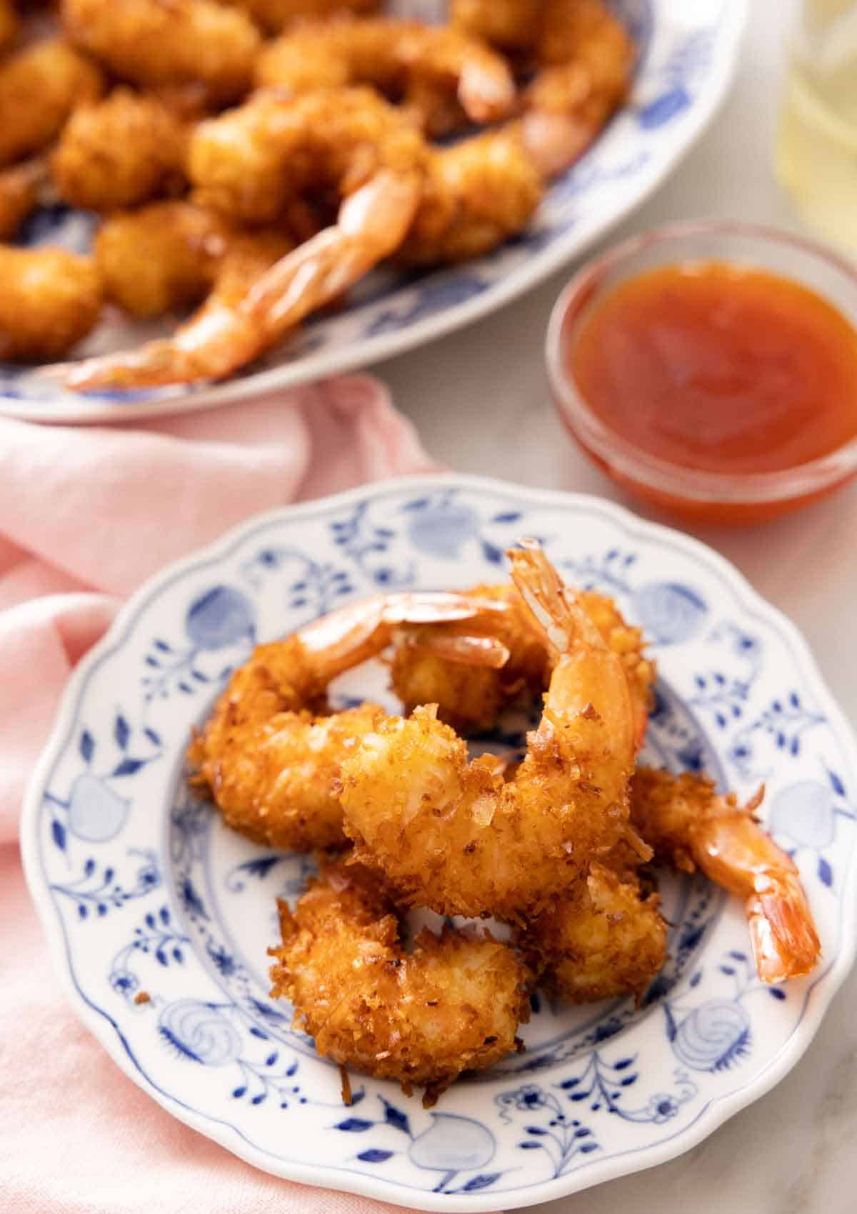 A small plate with five coconut shrimps in front of a dipping sauce and a platter with more shrimp.