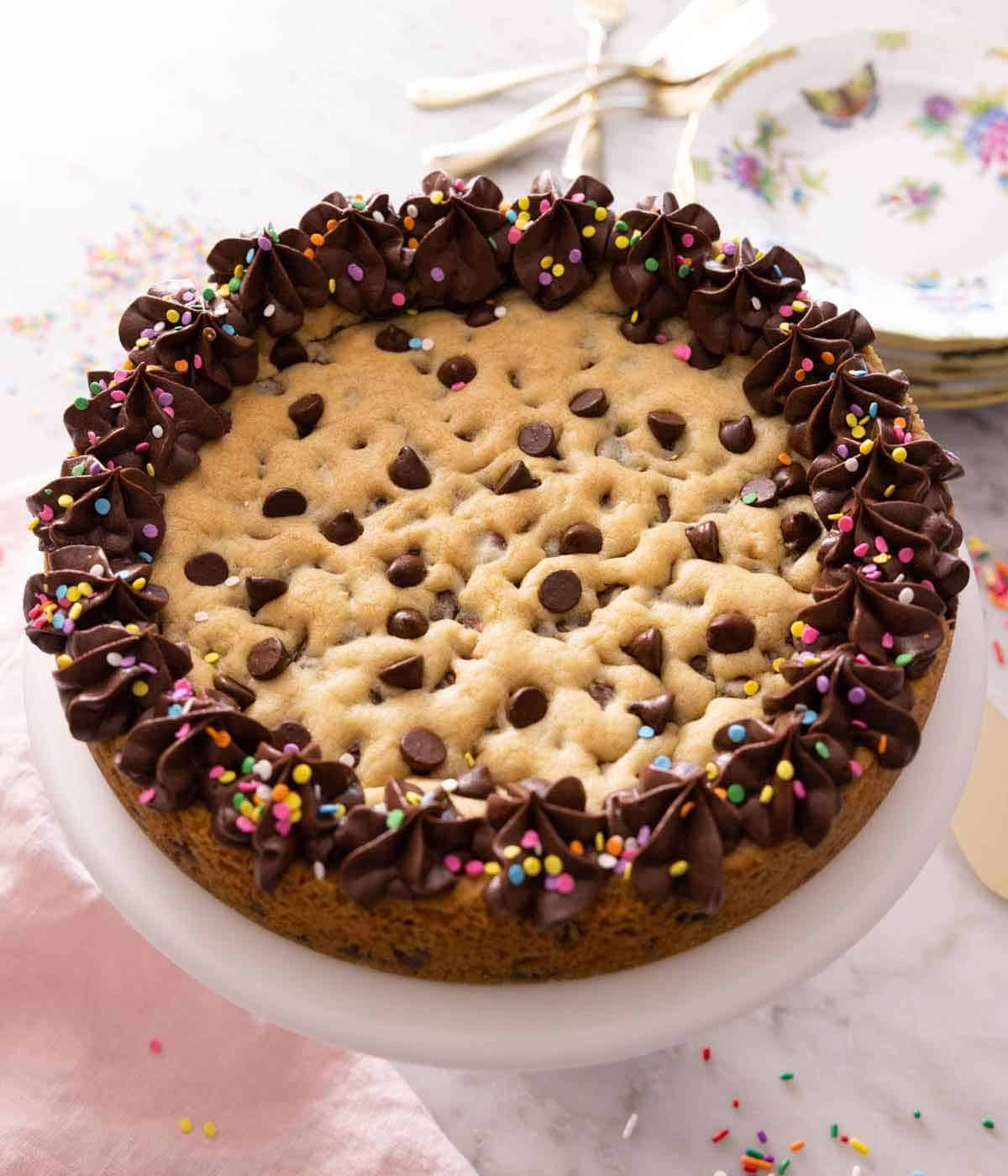 A cookie cake with chocolate frosting with sprinkles on a cake stand.