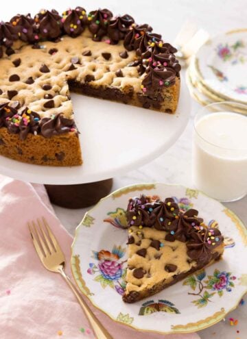 A cake stand with a cookie cake with a slice removed and placed on a plate.