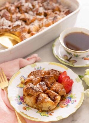 A plate with a serving of French toast casserole with sliced strawberries in front of a cup of coffee and a casserole dish.