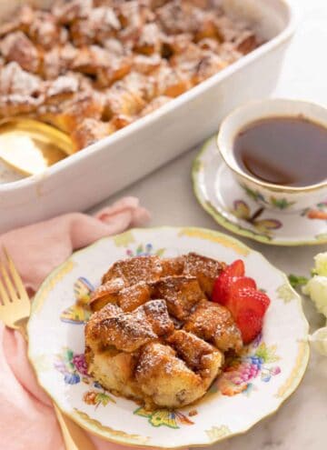 A plate with a serving of French toast casserole with sliced strawberries in front of a cup of coffee and a casserole dish.
