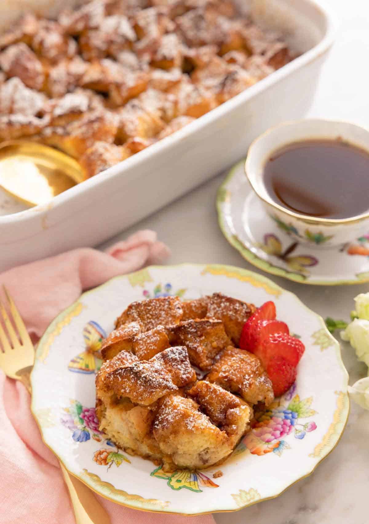 A plate with a serving of French toast casserole with sliced strawberries in front of a cup of coffee and a casserole dish.